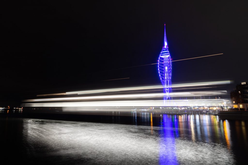 A purple Spinnaker Tower at night in Portsmouth Harbour with the white lights of a ferry streaking past