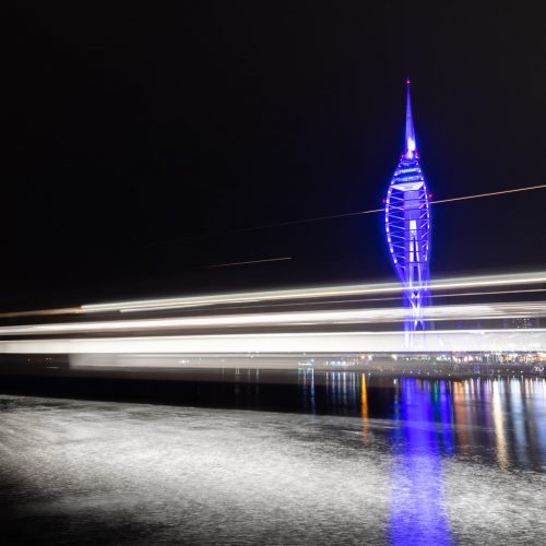 A purple Spinnaker Tower at night in Portsmouth Harbour with the white lights of a ferry streaking past